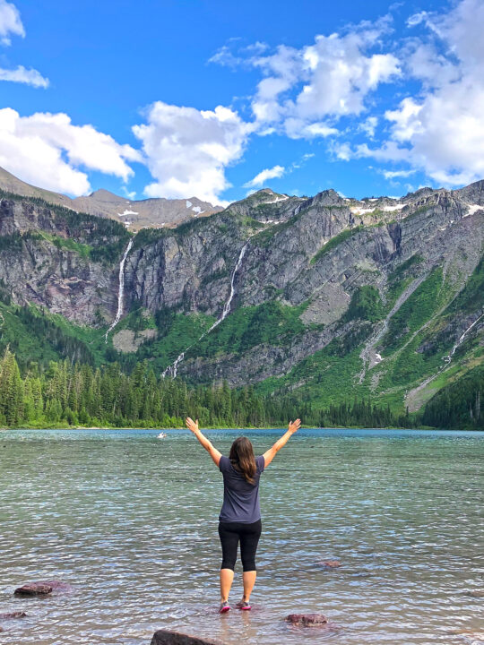 woman standing at Avalanche Lake with hands in the air with mountains and waterfalls in distance