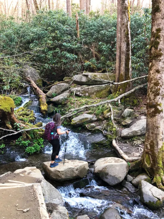 woman with hiking backpack stepping over large stones near waterfall with trees and shrubs nearby