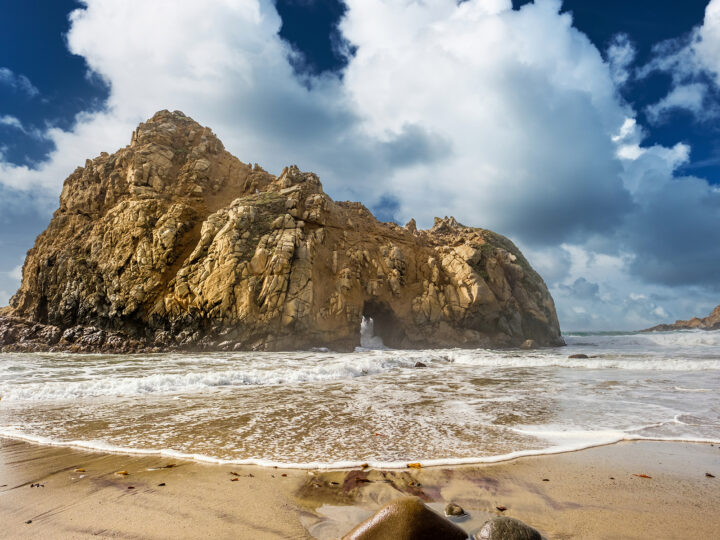 large rock on beach with waves and puffy clouds