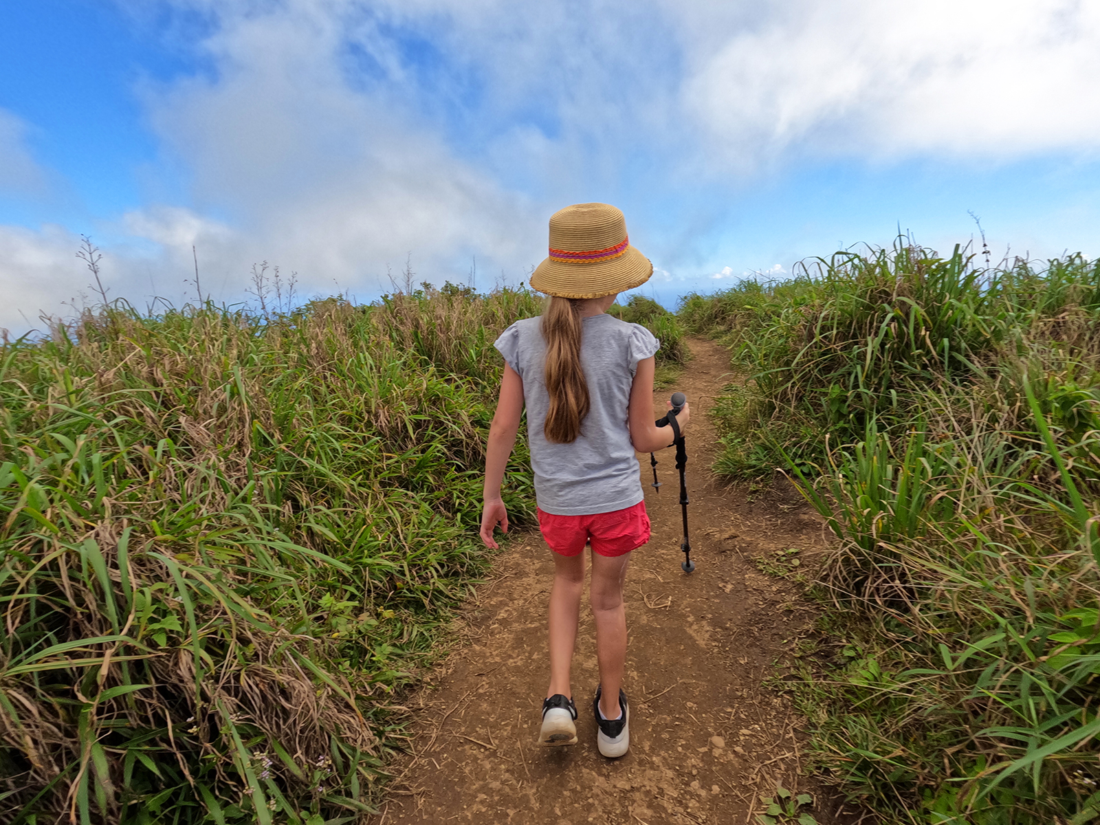 girl with long hair hat hiking on dirt path with tall grass surrounding her