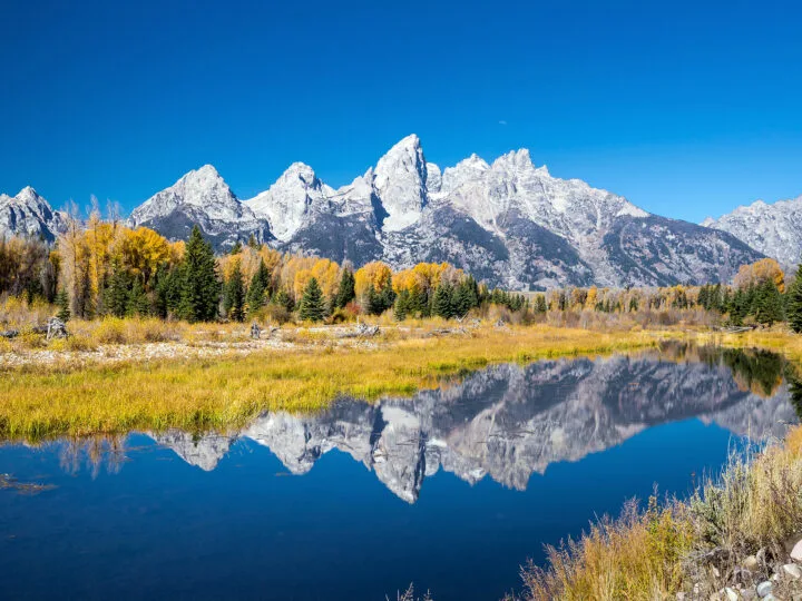 best national parks to visit in October white jagged mountain peaks fall foliage and lake reflection at Grand Teton