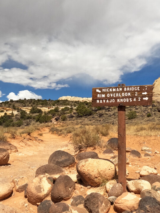 hickman bridge sign with rocks at base trail in distance on cloudy sky