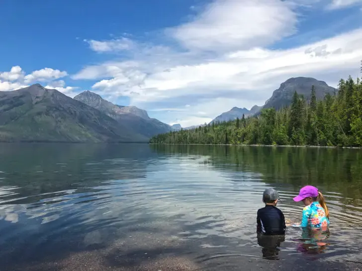 view of two kids sitting in water with lake scene and mountains
