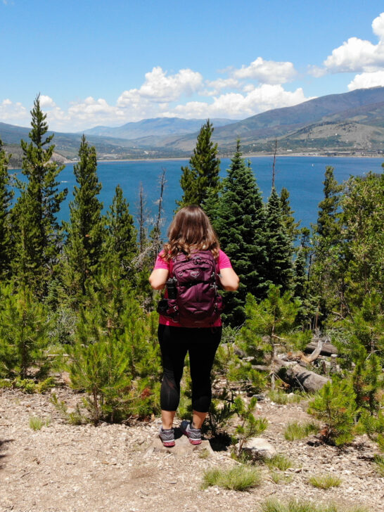 woman wearing hiking backpack standing near lake and trees