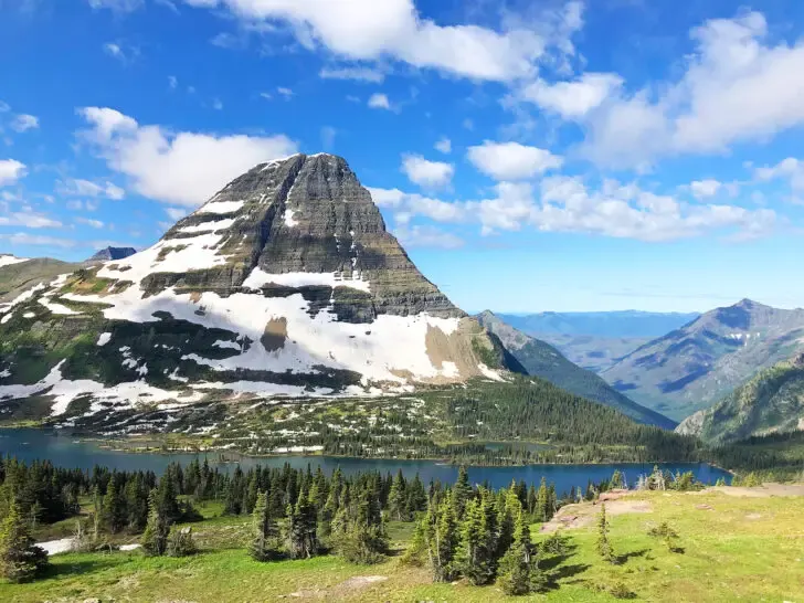 best places to visit in August view of mountain peak with snow and lake with trees and more mountains in the distance