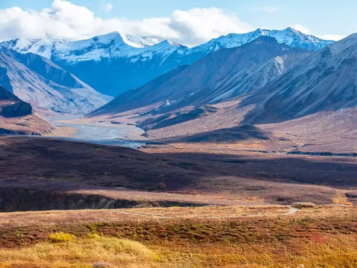 best place to vacation in August view of white capped mountains in Alaska with wide open field and road