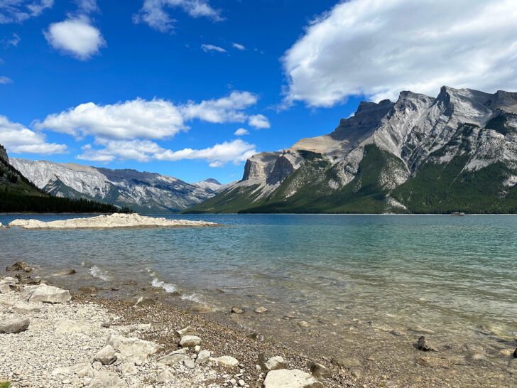 Banff summer scene with lake and rocky shore with jagged mountain peaks in distance