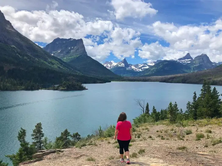woman hiking glacier national park with pink shirt black pants with lake and mountains in distance