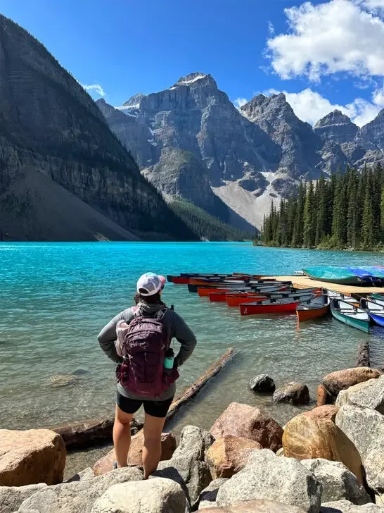 woman standing at lake with backpack on and canoes and mountains in distance
