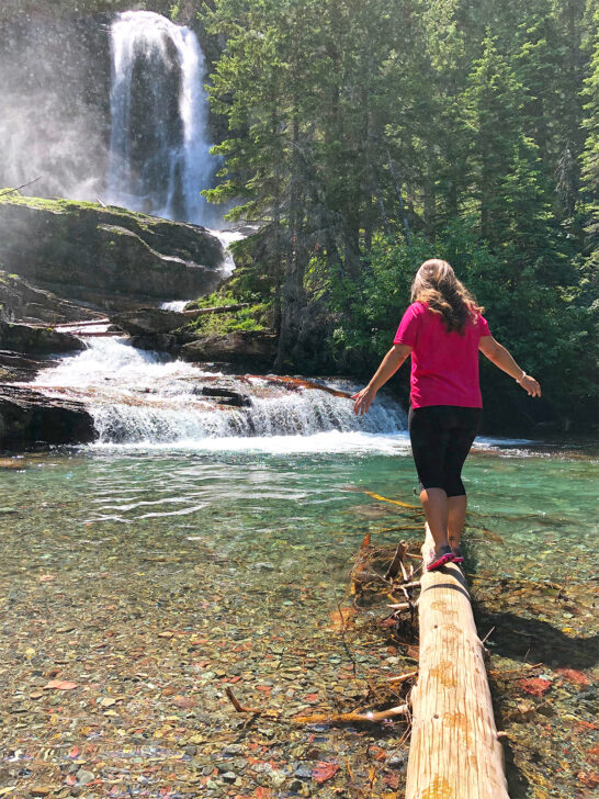 woman in pink shirt black pants standing on log in glacier national park