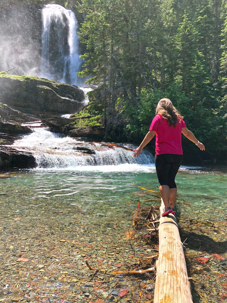 woman in pink shirt black pants standing on log in waterfall easy hikes glacier national park