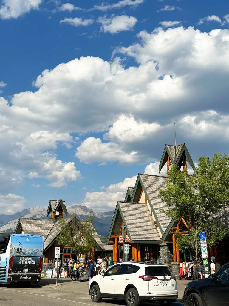 view of town with log cabin style stores and cars with mountains and puffy clouds in the sky