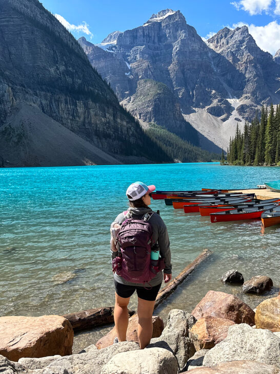 banff road trip view of woman standing by lake with bright blue water and canoes with mountains