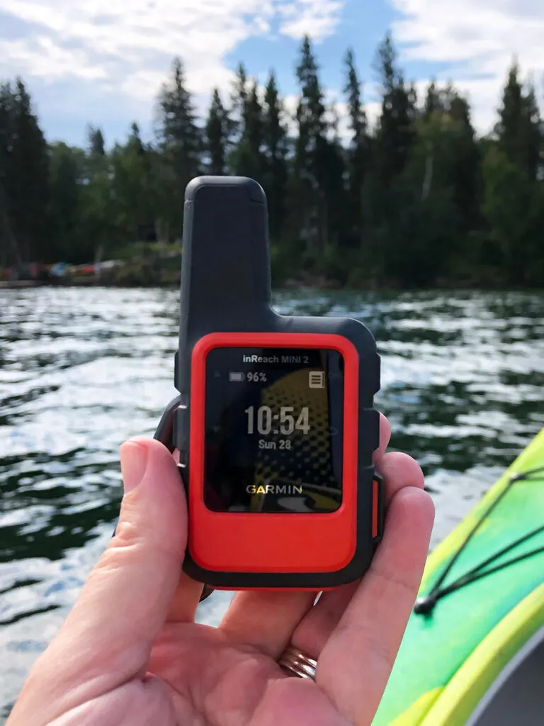 hand holding device that says Garmin inReach mini 2 10:54 Sun 28 with lake and kayak in distance
