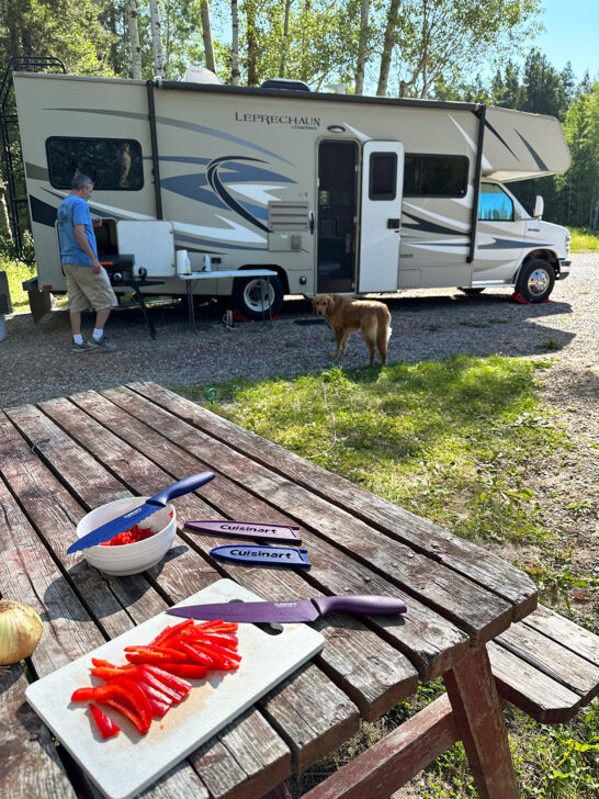 knives and cutting board with peppers on picnic table with camper in distance