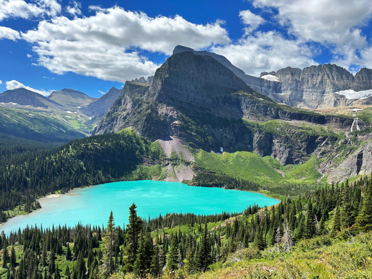 best lakes at glacier national park to visit view of teal colored lake with mountain peaks in background with trees surrounding