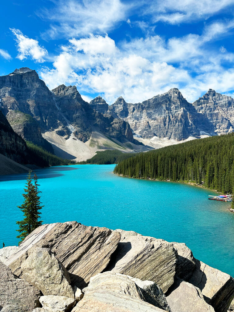 view of teal lake with rocky ledge and mountains in distance while on a road trip from Glacier to Banff